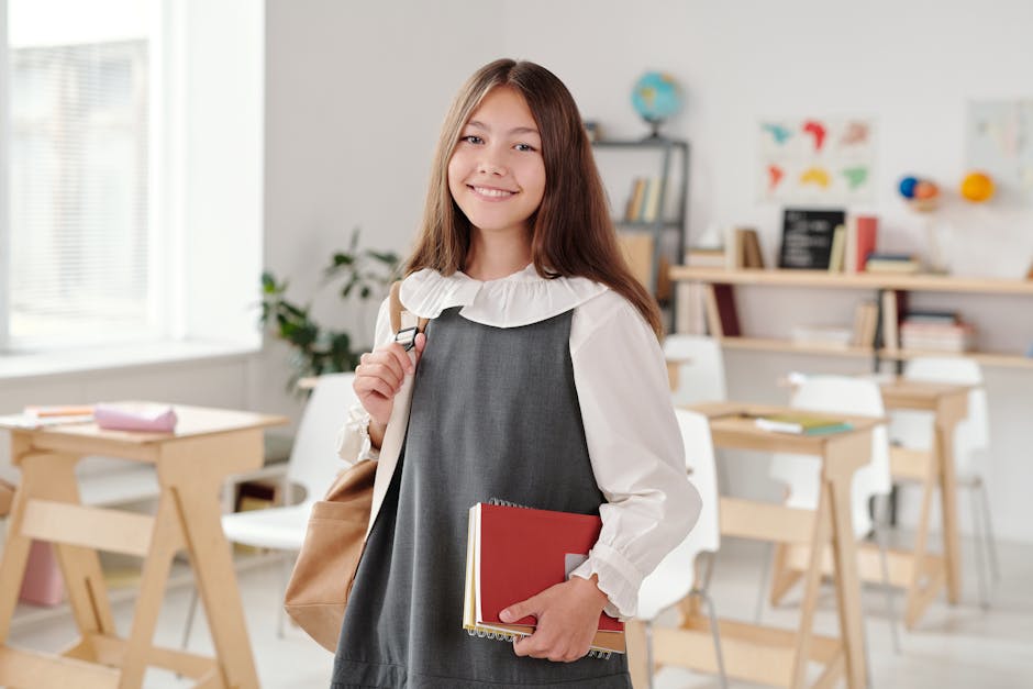 Smiling teenage girl in a school uniform holding notebooks in a bright classroom.